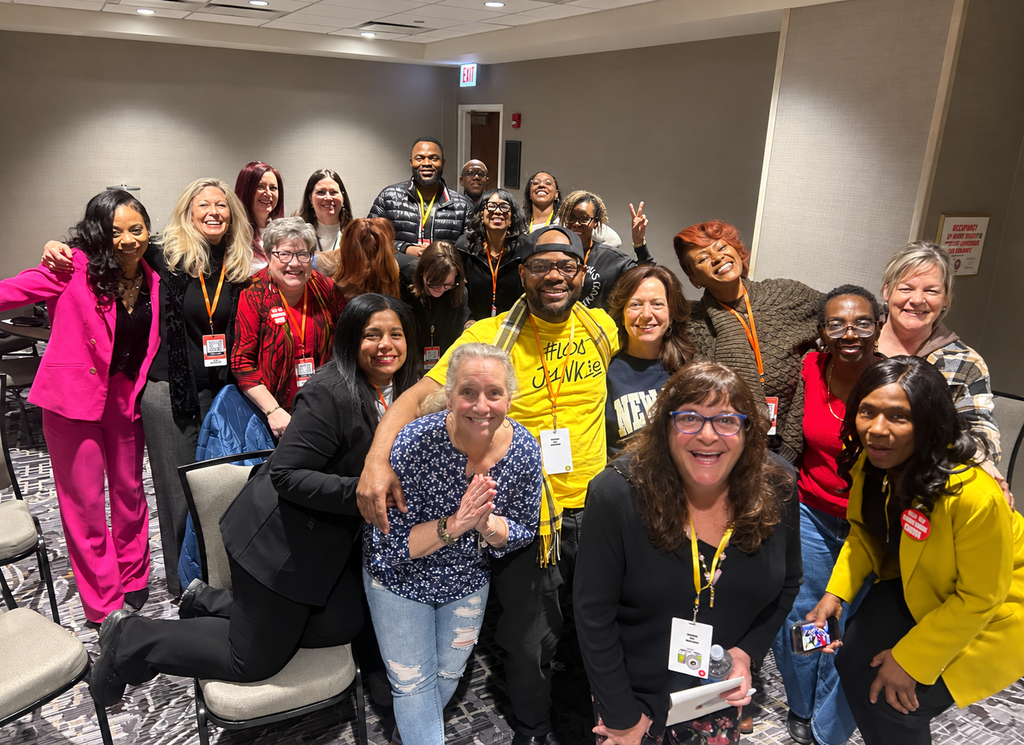 A smiling group of event attendees wearing lanyards gathered together with TM Hyman in a hotel meeting room.