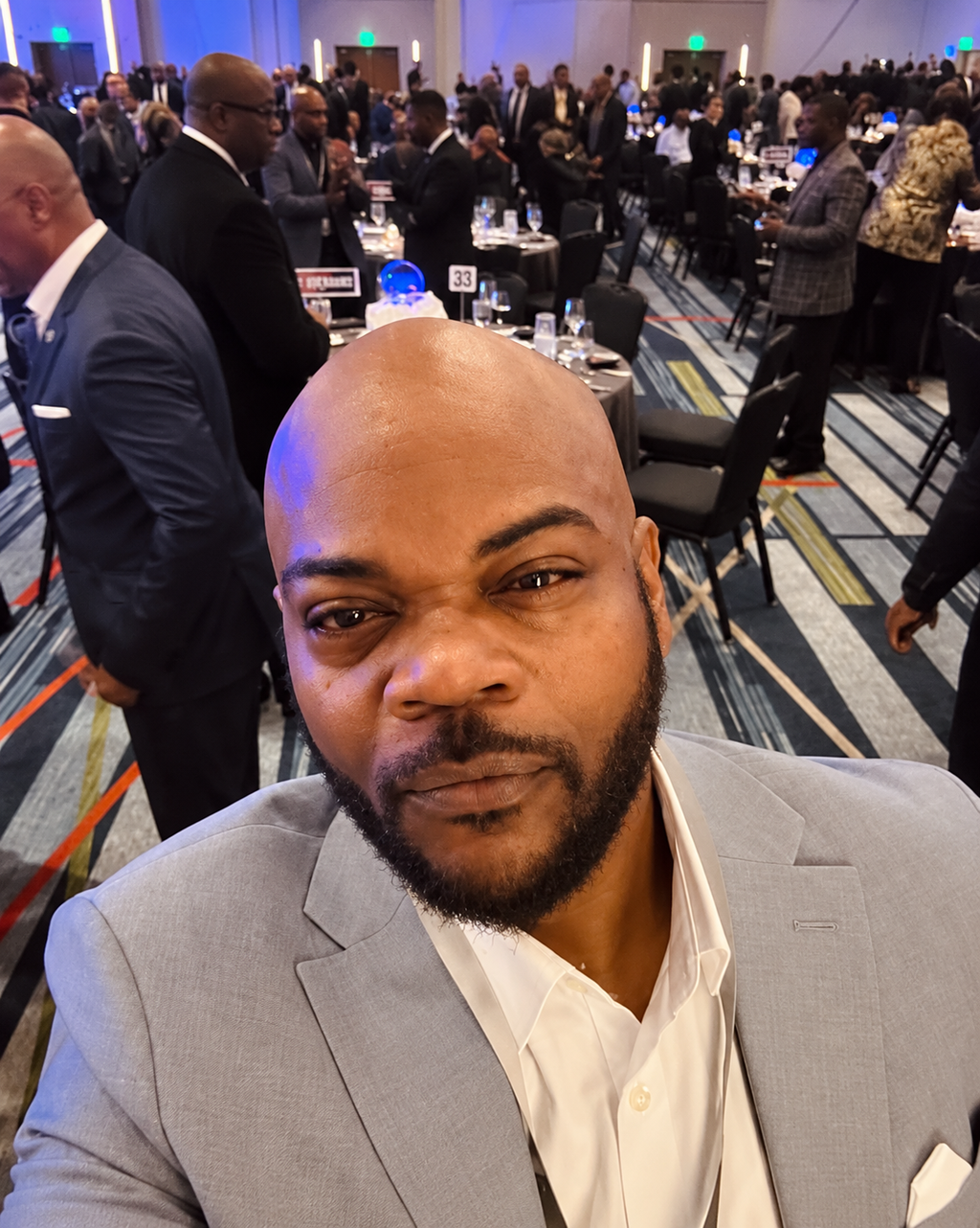 Portrait of TM Hyman in a light grey suit at a crowded conference dinner.