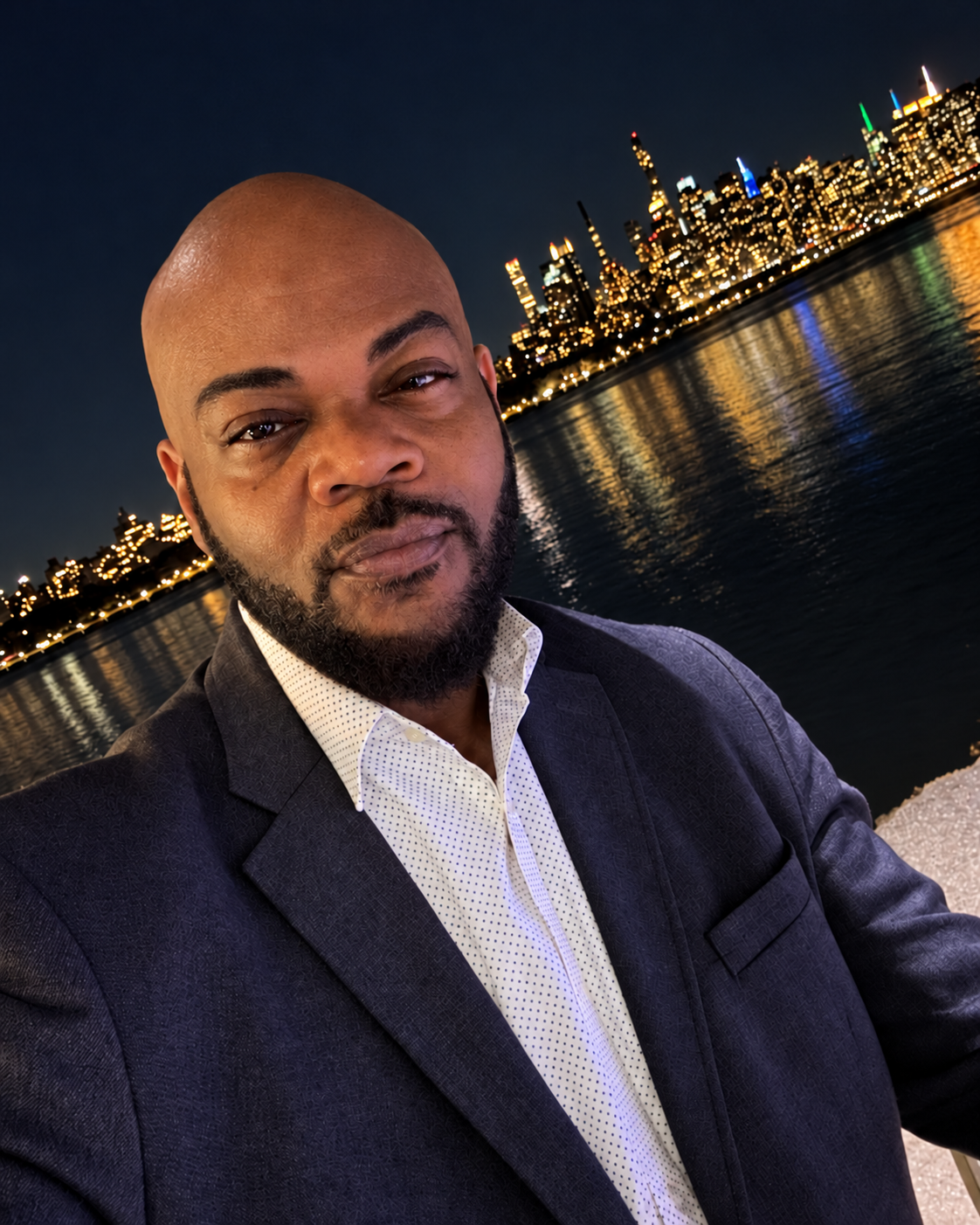 Portrait of TM Hyman at night in a dark blazer, with a lit city skyline reflected on the water behind him.