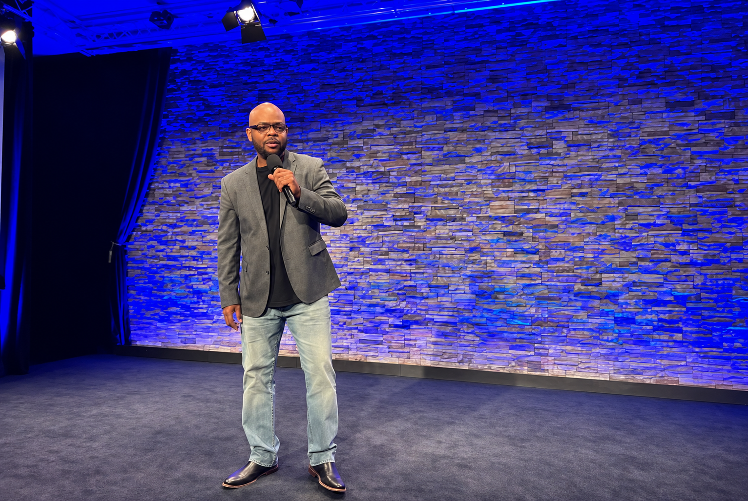 TM Hyman speaking into a microphone on a stage lit in deep blue, wearing a grey blazer and jeans.